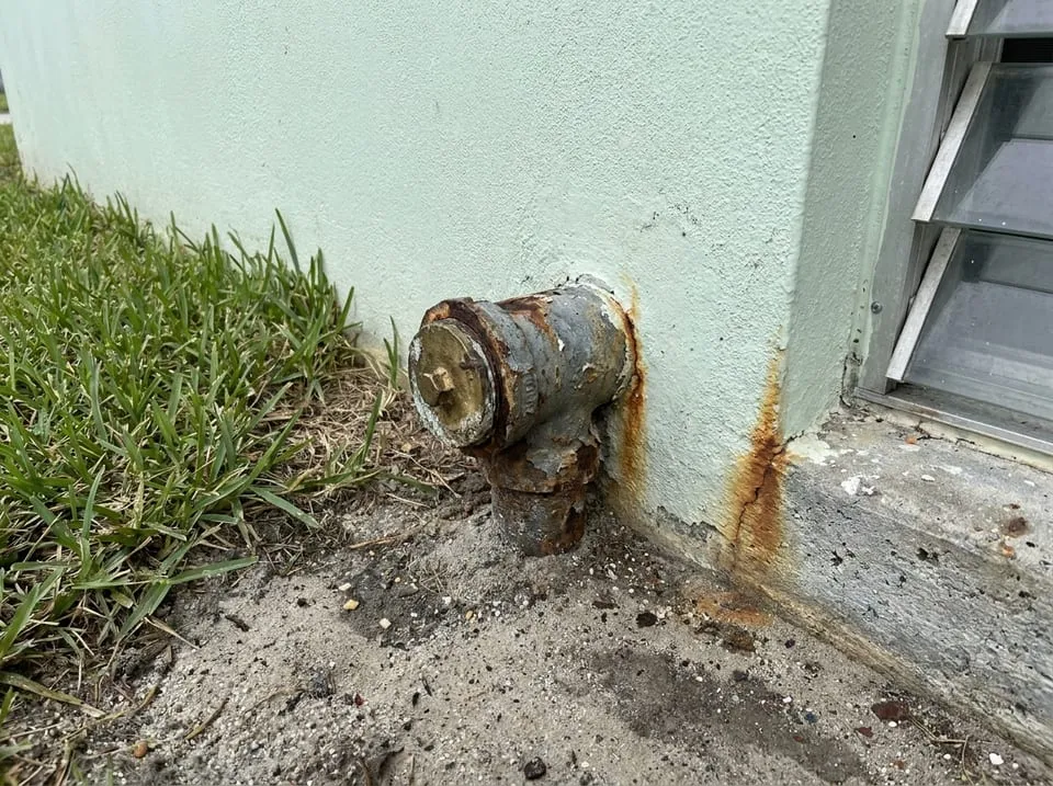Rusted cast iron cleanout at the stucco wall of a 1960s Florida house, a clue the home still has cast iron pipes