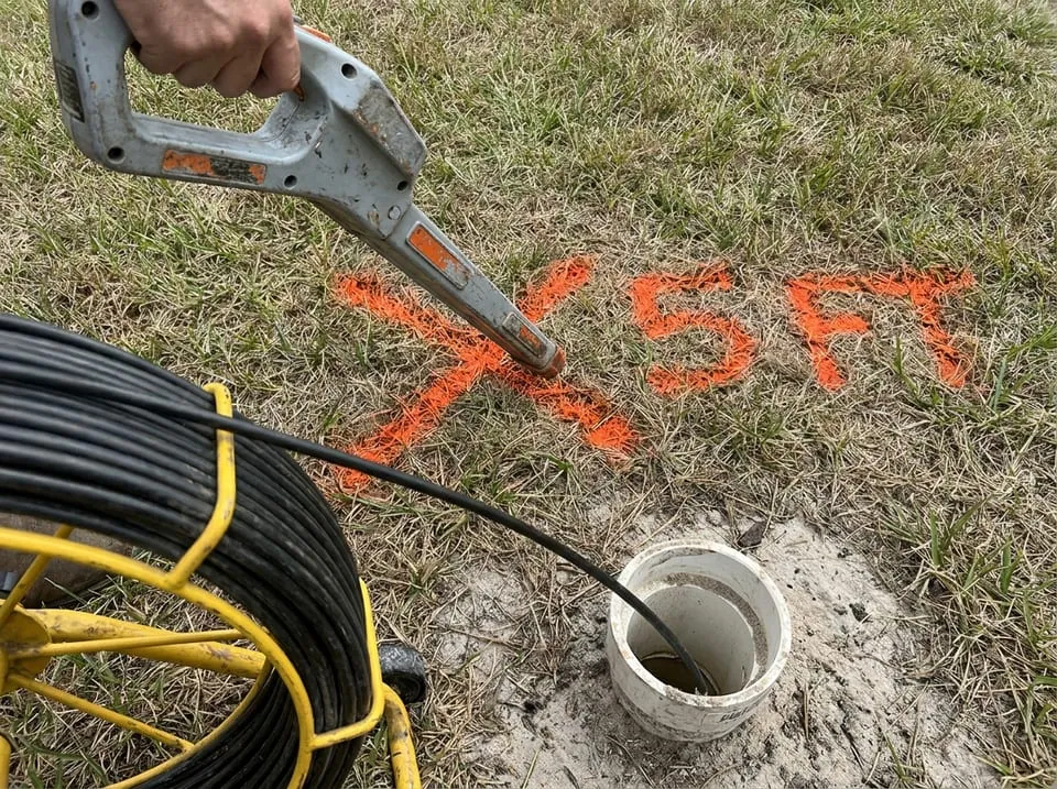 Pipe locator wand over a spray-paint mark and depth figure on a Florida lawn during a sewer camera inspection